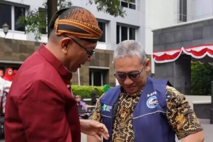 Two men in an outdoor plaza share a moment, one in a maroon shirt and patterned headwrap, the other in a blue vest and batik shirt, looking at the other's hand.