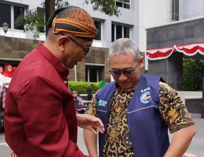 Two men in an outdoor plaza share a moment, one in a maroon shirt and patterned headwrap, the other in a blue vest and batik shirt, looking at the other's hand.