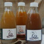Three glass bottles with white caps containing brown liquid sit on a wooden surface; front labels show a person and the word 'Jog Jam'.