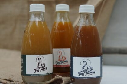 Three glass bottles with white caps containing brown liquid sit on a wooden surface; front labels show a person and the word 'Jog Jam'.