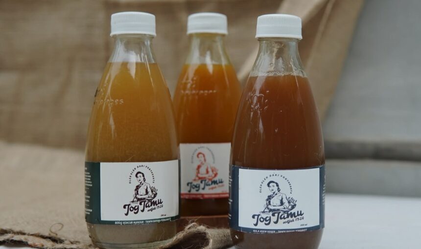 Three glass bottles with white caps containing brown liquid sit on a wooden surface; front labels show a person and the word 'Jog Jam'.