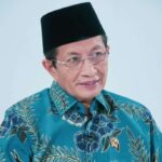 Older man wearing a black peci hat and glasses, in a turquoise floral batik shirt, looking to the side against a light background.