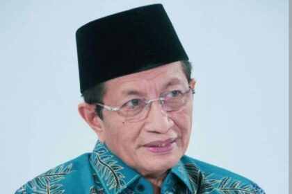 Older man wearing a black peci hat and glasses, in a turquoise floral batik shirt, looking to the side against a light background.