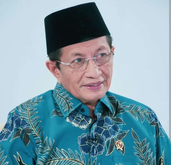 Older man wearing a black peci hat and glasses, in a turquoise floral batik shirt, looking to the side against a light background.