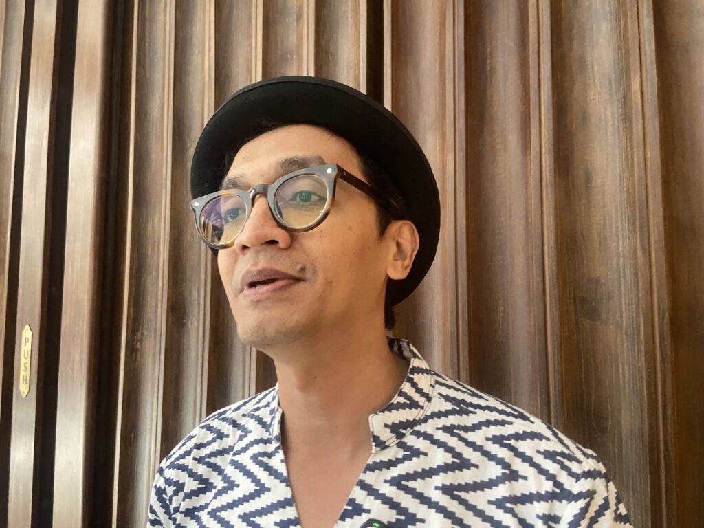 IMG_0017 - Mabur.co Portrait of a person wearing a black hat and round glasses, standing in front of vertical wood panels.
