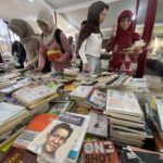 Women in hijabs browse a crowded book stall with tables piled with books inside a tent-like market area.
