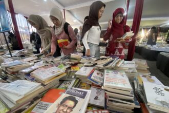 Women in hijabs browse a crowded book stall with tables piled with books inside a tent-like market area.