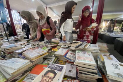 Women in hijabs browse a crowded book stall with tables piled with books inside a tent-like market area.
