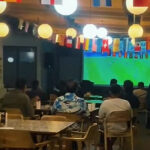 Customers seated at tables in a cafe watching a large soccer match on a wall screen, colorful flags hanging overhead.