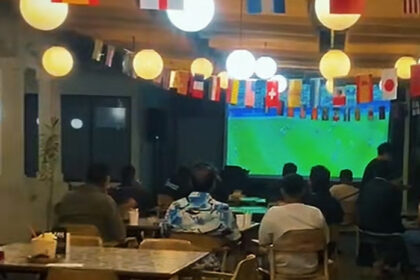Customers seated at tables in a cafe watching a large soccer match on a wall screen, colorful flags hanging overhead.