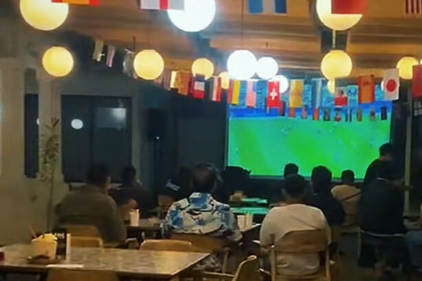 Customers seated at tables in a cafe watching a large soccer match on a wall screen, colorful flags hanging overhead.