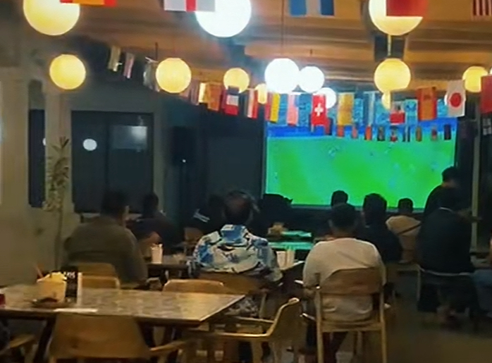 Customers seated at tables in a cafe watching a large soccer match on a wall screen, colorful flags hanging overhead.