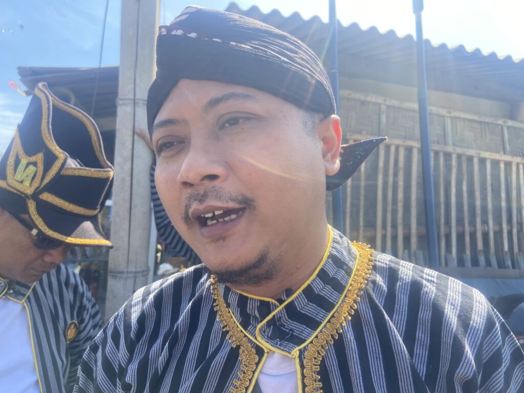 Selfie of a man in a striped ceremonial jacket with gold trim, wearing a black headwrap, outdoors with a wooden fence in the background.