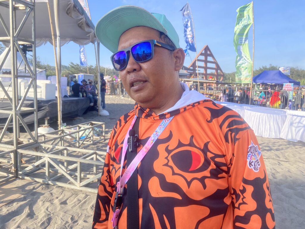 Man in orange tiger-pattern shirt with blue mirrored sunglasses at an outdoor festival, with stage scaffolding behind.