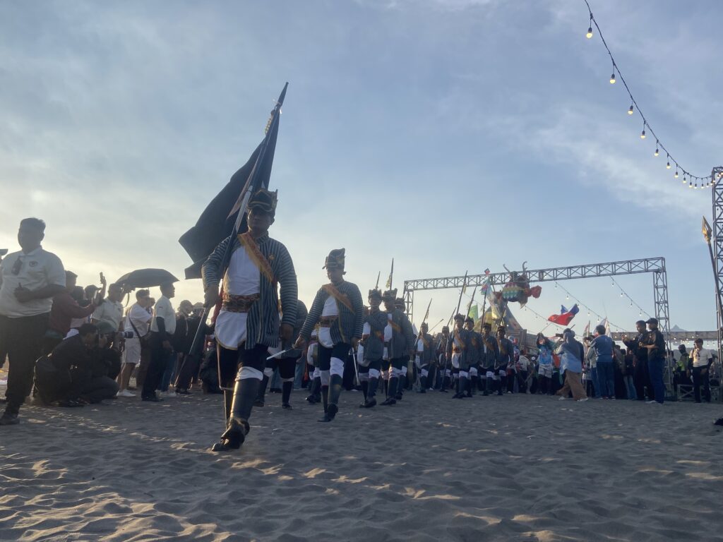 Group of uniformed performers marching on a sandy beach during a parade, flags raised, with spectators lining the edge and string lights above.
