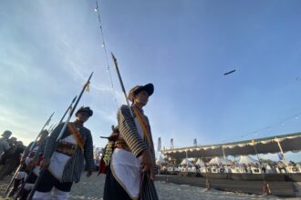 Men in traditional striped costumes marching with spears during an outdoor festival by the beach, with a crowd seated at a decorated stage and string lights overhead.