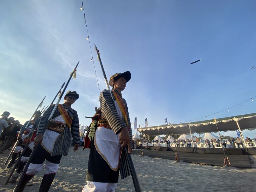 Men in traditional striped costumes marching with spears during an outdoor festival by the beach, with a crowd seated at a decorated stage and string lights overhead.