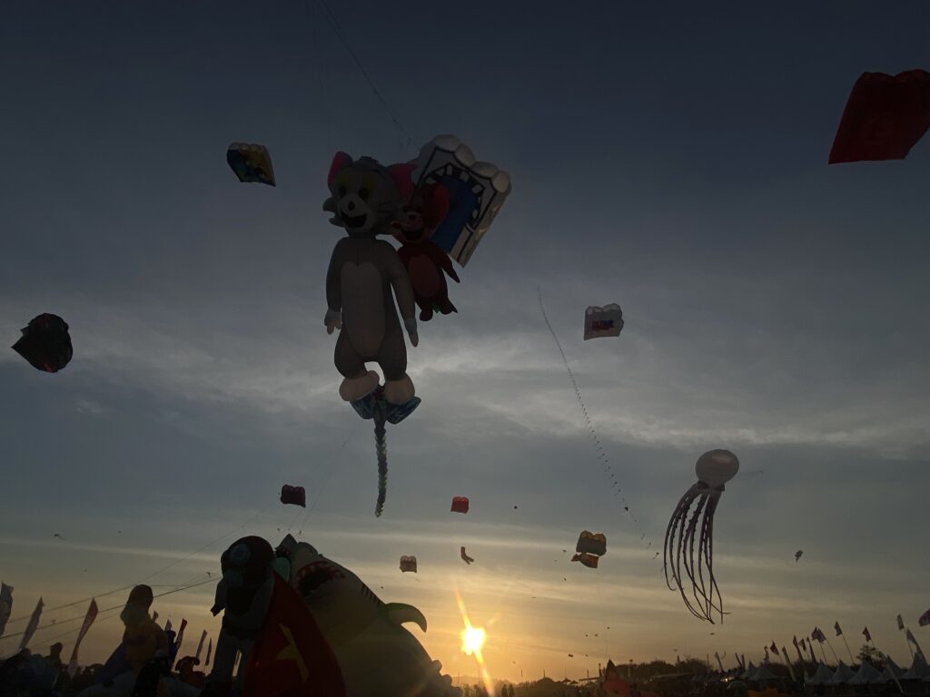 Giant cartoon character kite with other colorful kites flying in a dusk sky at a festival.
