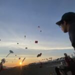 Kite festival at sunset with numerous colorful kites in the sky and people gathered along the beach.