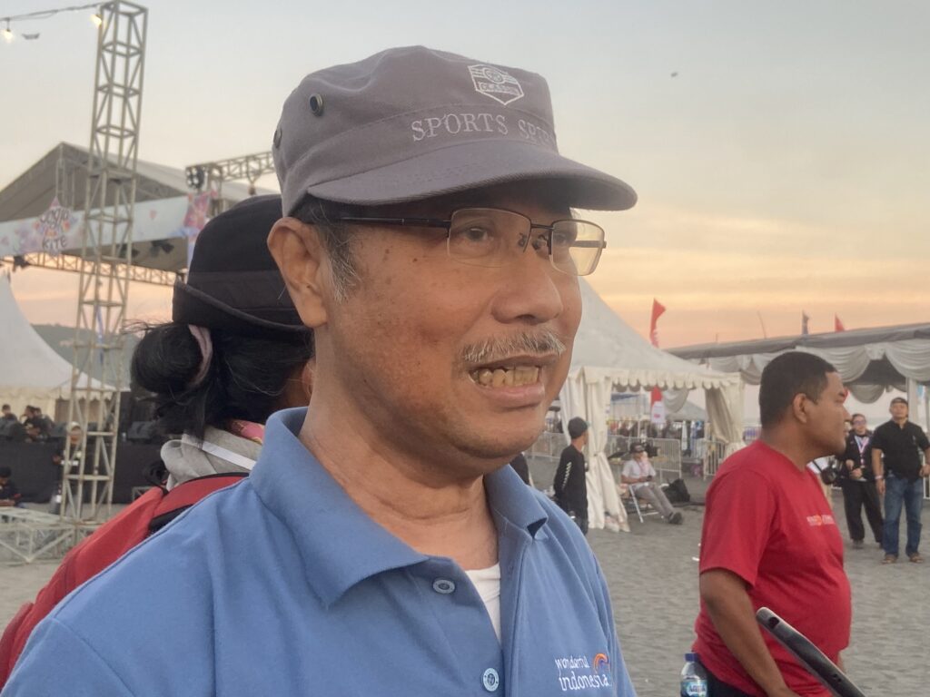 Man wearing a gray hat and blue polo shirt at an outdoor festival, tents and a stage structure visible in the background.