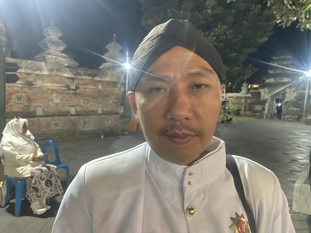 Selfie of a man in a white traditional outfit with gold buttons, night-time temple walls in the background.