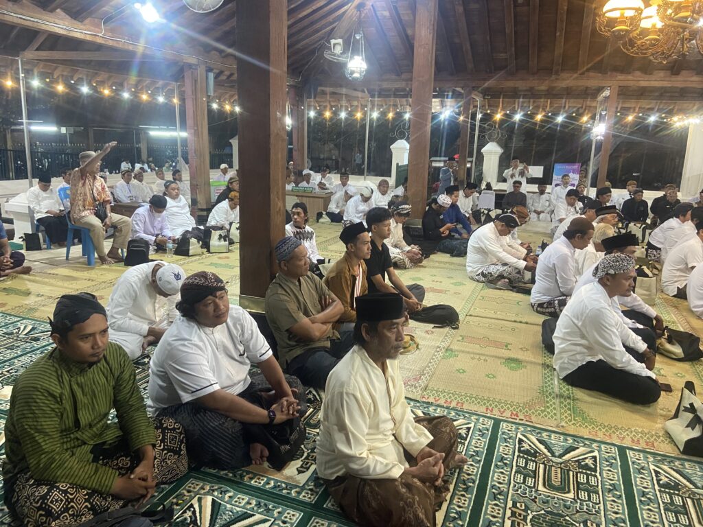 Group of men in white and traditional clothing seated on prayer mats inside a wooden pavilion with string lights overhead, likely a religious gathering at night.