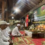 Group of people in white traditional clothing seated on a patterned carpet inside a wooden hall, with a green banner, flags, and floral arrangements at a ceremonial event.