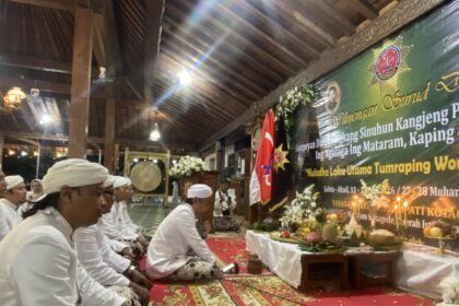 Group of people in white traditional clothing seated on a patterned carpet inside a wooden hall, with a green banner, flags, and floral arrangements at a ceremonial event.