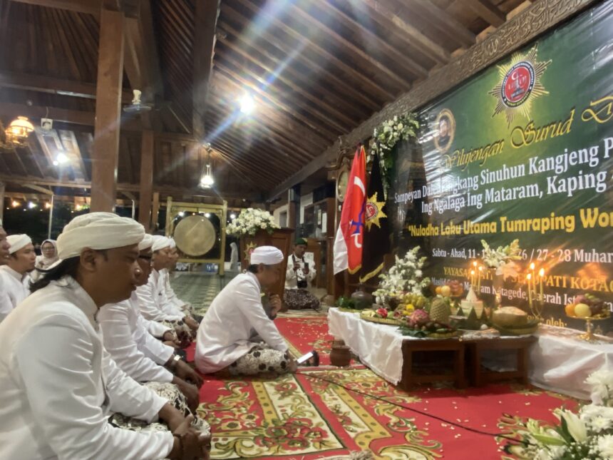Group of people in white traditional clothing seated on a patterned carpet inside a wooden hall, with a green banner, flags, and floral arrangements at a ceremonial event.