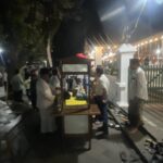 Nighttime outdoor party with string lights, people mingling near market stalls and a white fence in the background.