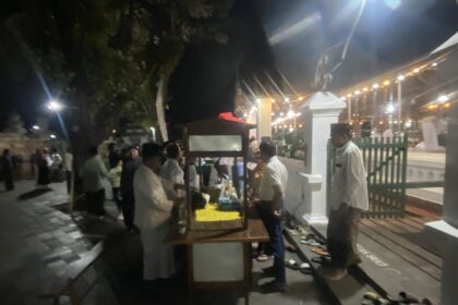 Nighttime outdoor party with string lights, people mingling near market stalls and a white fence in the background.