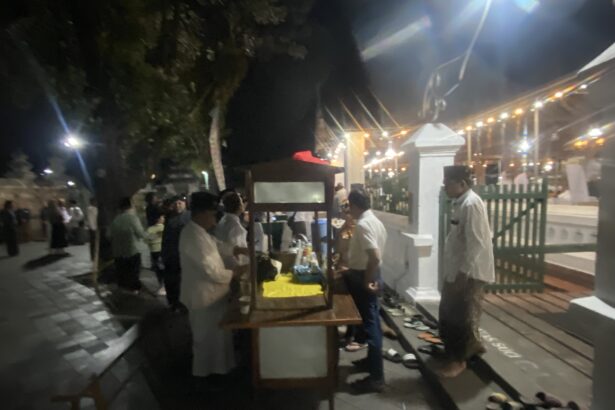 Nighttime outdoor party with string lights, people mingling near market stalls and a white fence in the background.