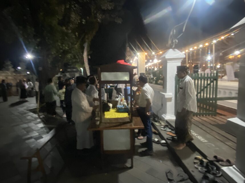 Nighttime outdoor party with string lights, people mingling near market stalls and a white fence in the background.