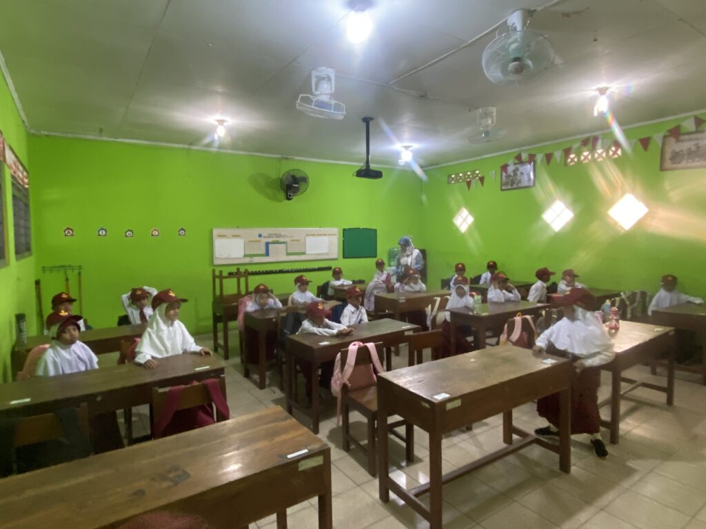 IMG_0270 - Mabur.co Classroom with green walls, students in white uniforms and maroon hats seated at wooden desks facing forward, a teacher at the front.