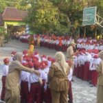Large group of schoolchildren in white shirts, red caps and maroon skirts/pants standing in rows for an outdoor assembly, supervised by teachers in tan uniforms.