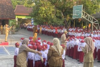 Large group of schoolchildren in white shirts, red caps and maroon skirts/pants standing in rows for an outdoor assembly, supervised by teachers in tan uniforms.