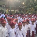 Crowd of schoolchildren in white uniforms and maroon hats gathered outdoors under a shaded schoolyard with trees nearby.