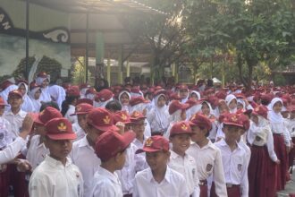 Crowd of schoolchildren in white uniforms and maroon hats gathered outdoors under a shaded schoolyard with trees nearby.