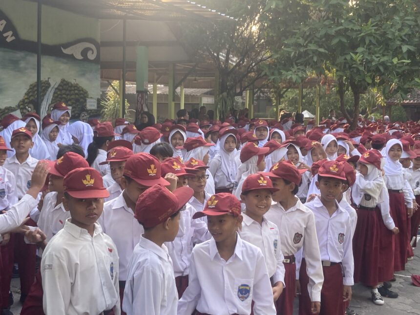 Crowd of schoolchildren in white uniforms and maroon hats gathered outdoors under a shaded schoolyard with trees nearby.