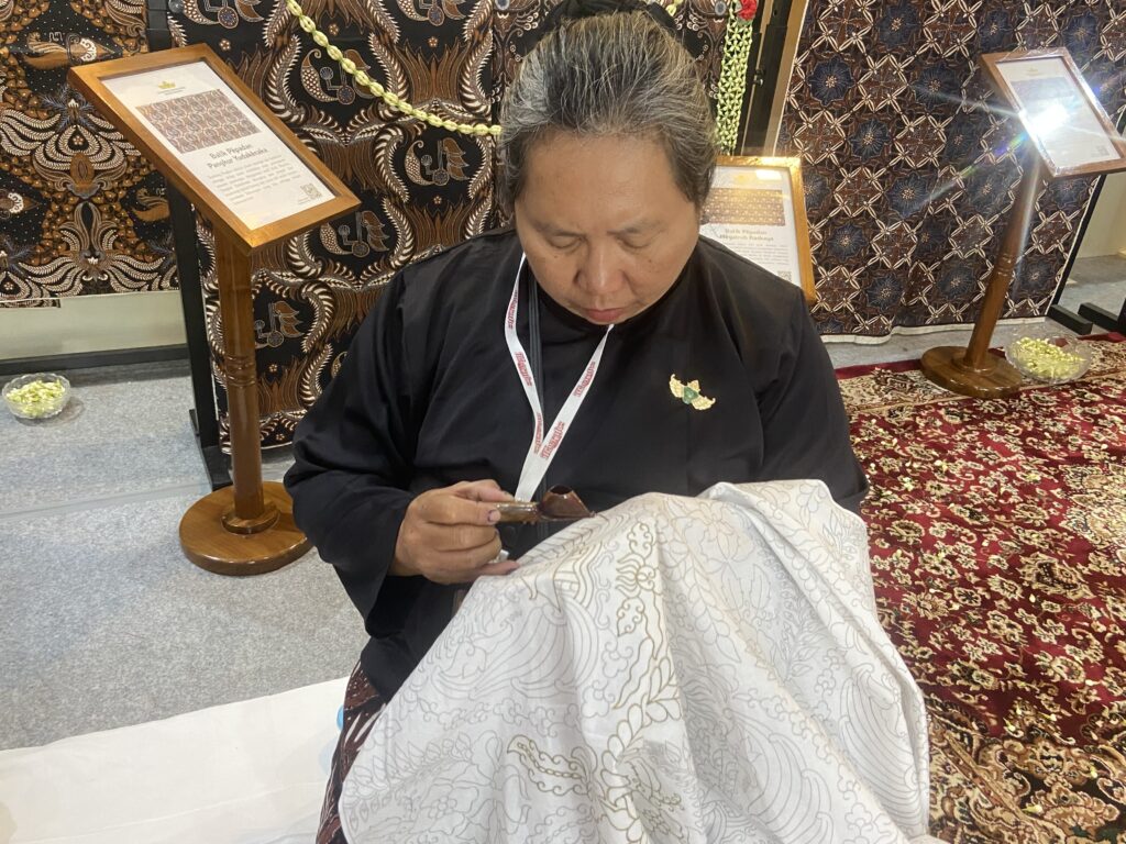 Person in a black shirt with a lanyard, seated and looking down as they hold a chocolate piece over a light patterned cloth in a decorative display area with batik fabric backdrop.