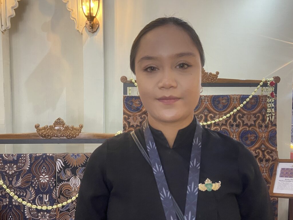 Close-up portrait of a woman in a black outfit with a blue lanyard, ornate batik background behind her.