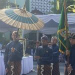 Group of men in matching blue batik outfits carry a large decorative parasol at an outdoor ceremonial setup with white-draped tents and greenery behind them.