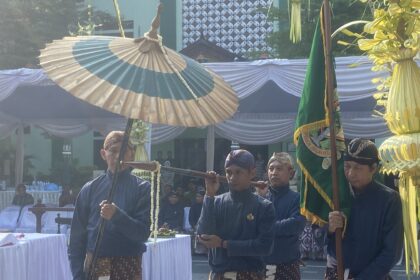 Group of men in matching blue batik outfits carry a large decorative parasol at an outdoor ceremonial setup with white-draped tents and greenery behind them.