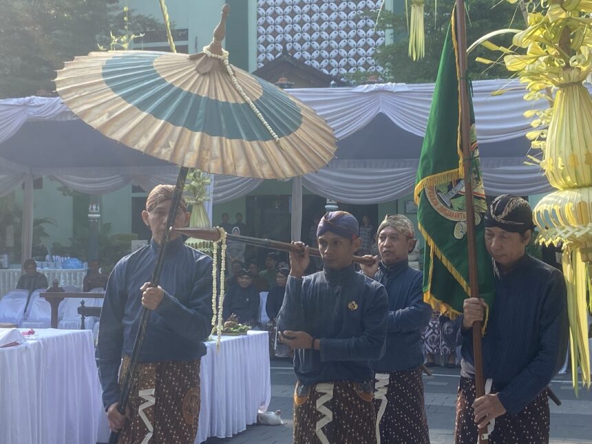 Group of men in matching blue batik outfits carry a large decorative parasol at an outdoor ceremonial setup with white-draped tents and greenery behind them.