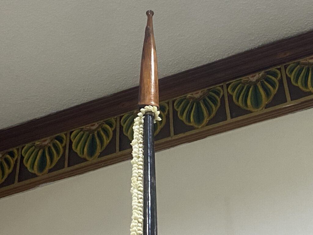 Ceremonial wooden spear with a pointed tip and a white flower garland hung indoors.