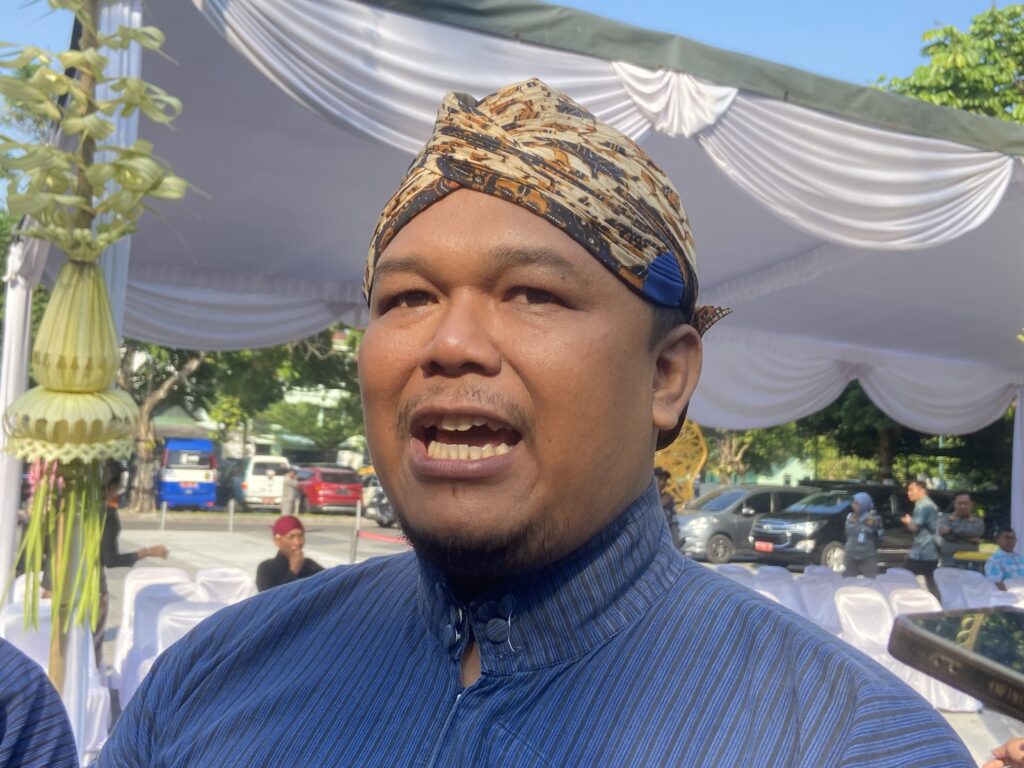 Close-up of a man in a patterned headwrap at an outdoor event with white drapery in the background.