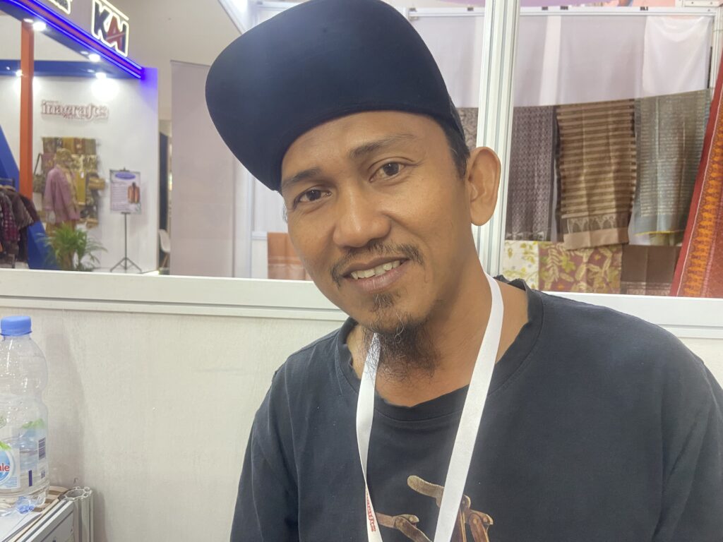 IMG_0456 1 - Mabur.co Close-up of a smiling man wearing a black cap in a showroom with rolled fabrics in the background behind him