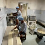Factory workers in masks and hairnets roll dough on a long table beside a pasta machine in a food processing plant.