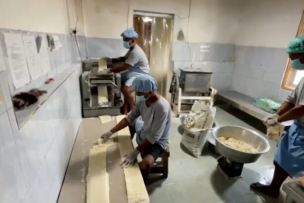 Factory workers in masks and hairnets roll dough on a long table beside a pasta machine in a food processing plant.