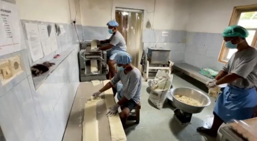 Factory workers in masks and hairnets roll dough on a long table beside a pasta machine in a food processing plant.